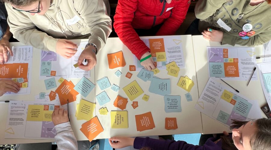 an overhead view of a table with various coloured text cards, sticky notes and pens. 7 people sit around the table, their hands reach for the cards on the table